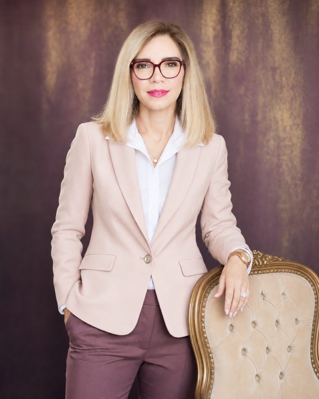 Blush blazer, burgundy frames. Standing, hand resting on a gilt armchair.