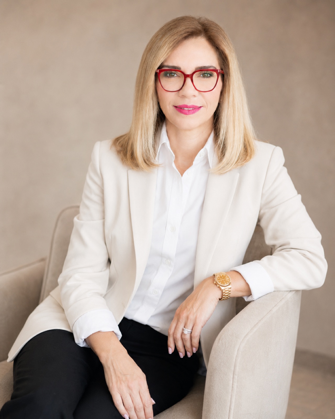 Ivory blazer, red frames. Seated in a cream armchair.