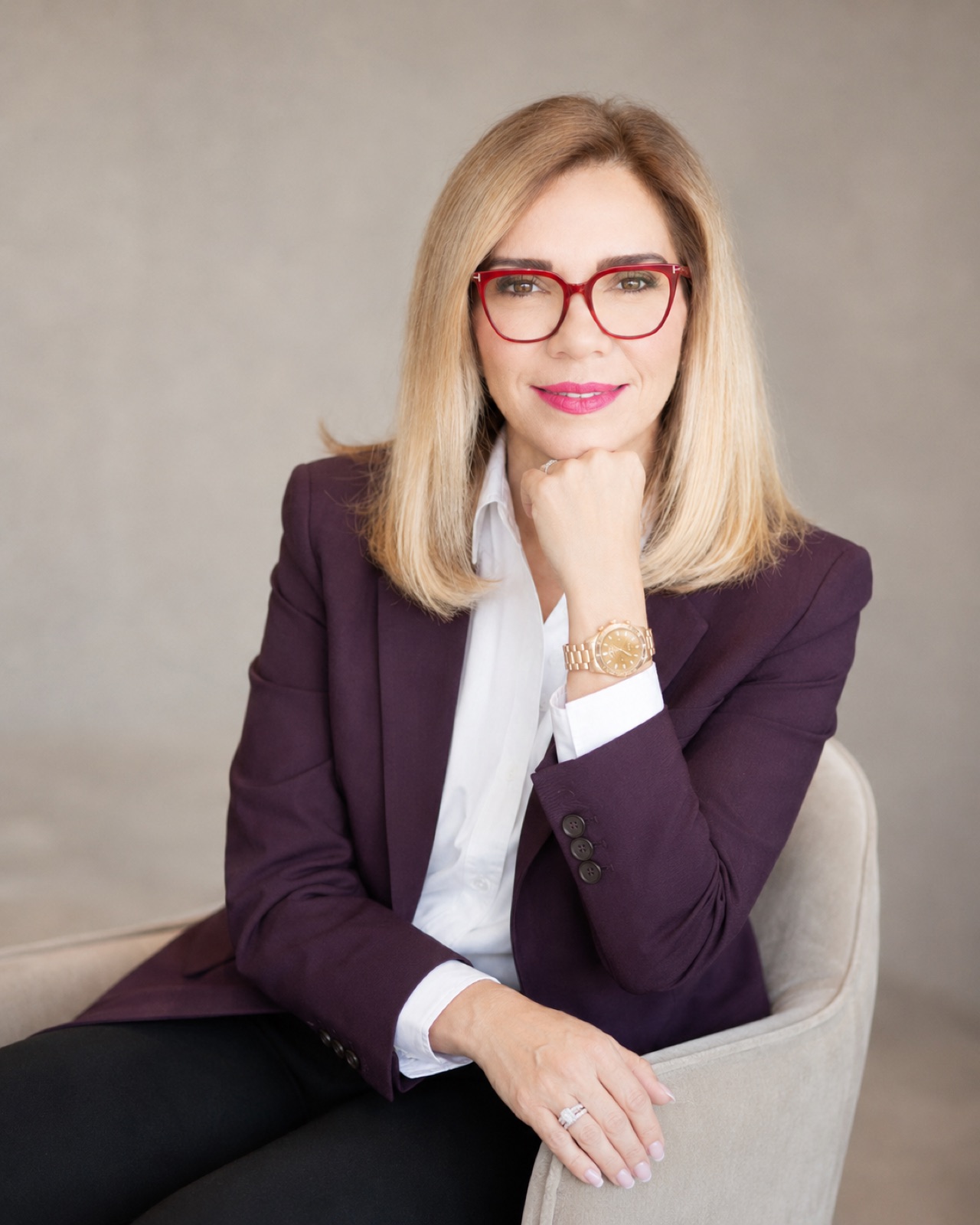 Plum blazer, red frames. Hand to chin, cream armchair.