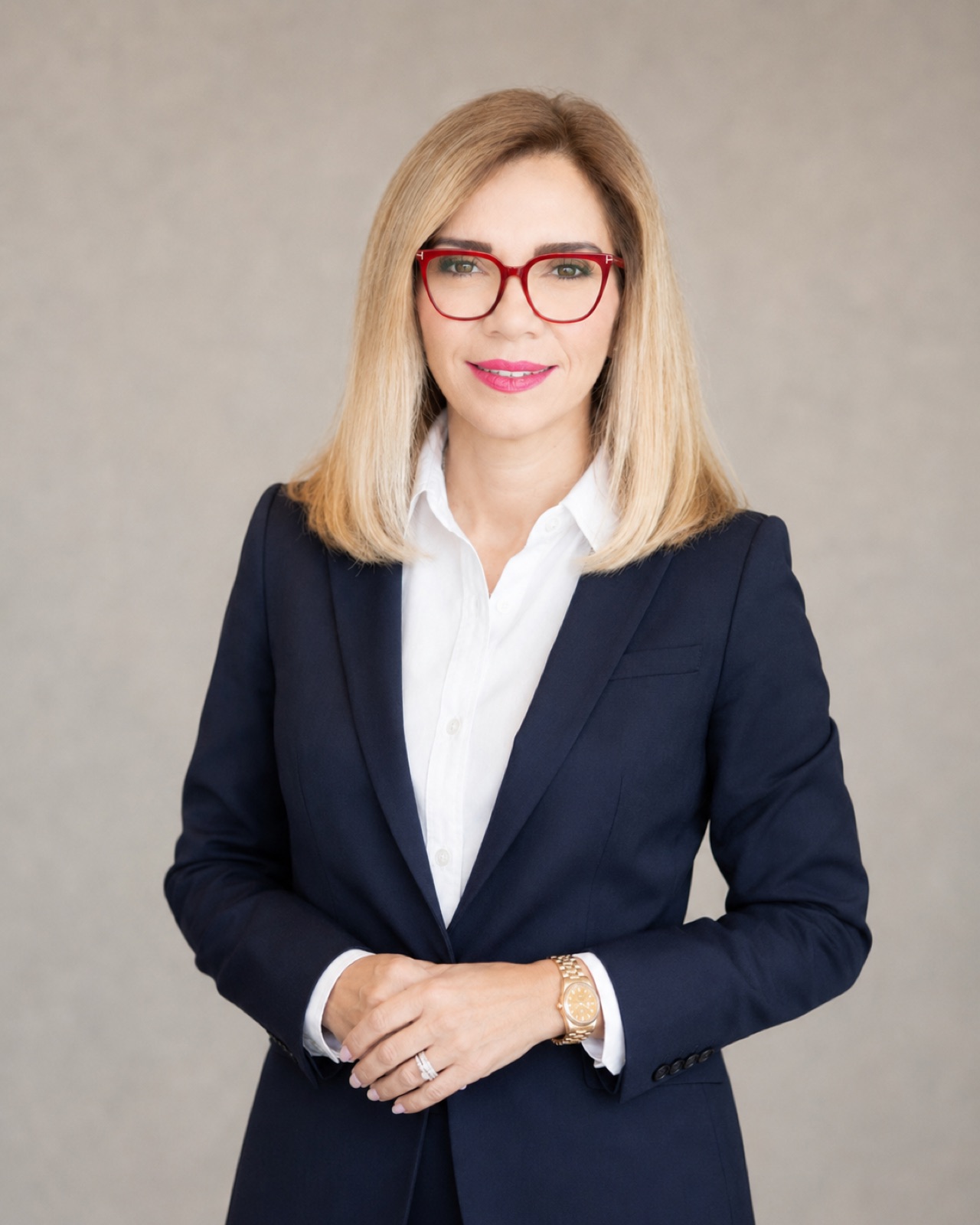 Navy suit, red frames. Standing, hands clasped, pale ground.