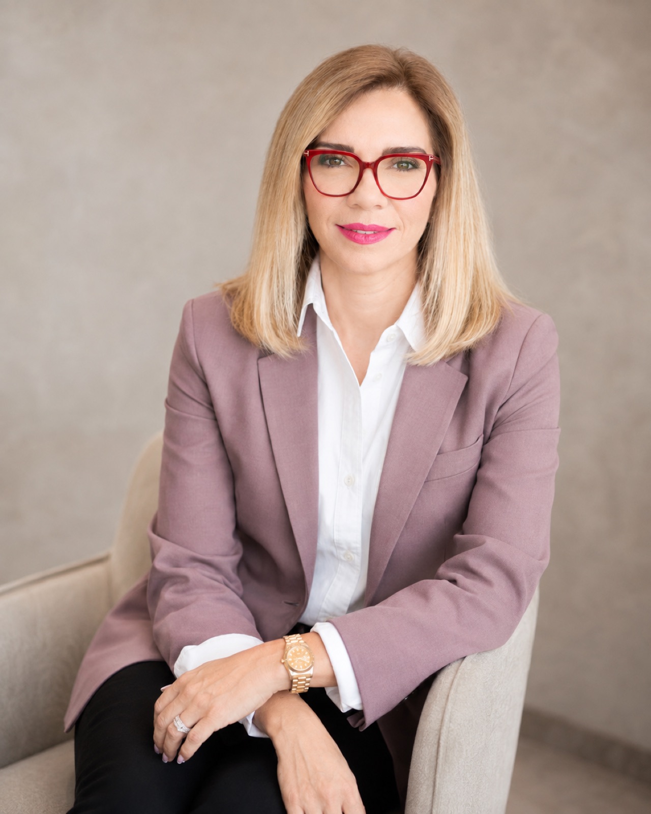Mauve blazer, red frames. Seated, pale ground.