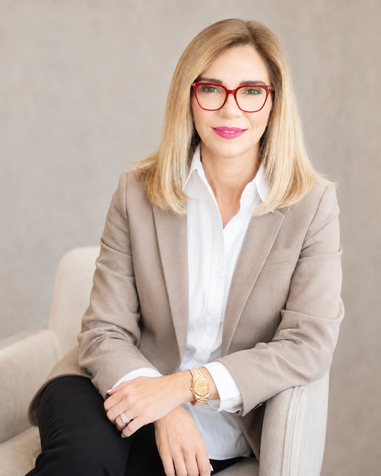 Taupe blazer, red frames. Seated in a cream armchair.