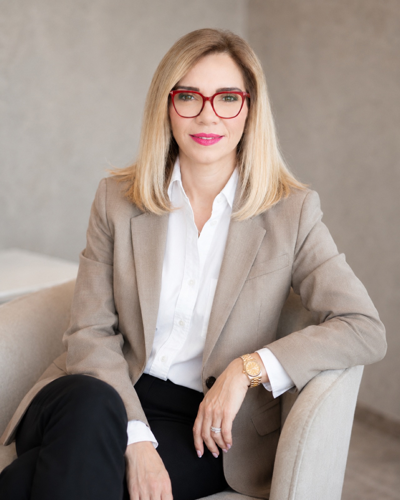 Taupe blazer, red frames. Seated, legs crossed, arm along the chair.