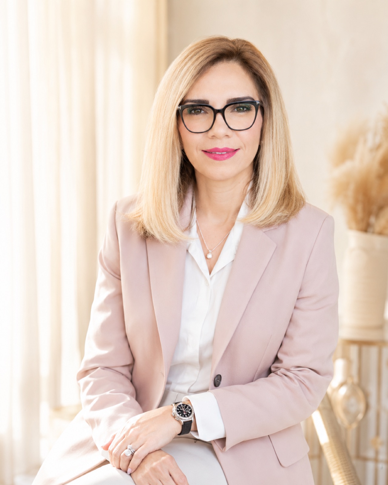Blush blazer, black frames. Bright window light, pampas grass behind.