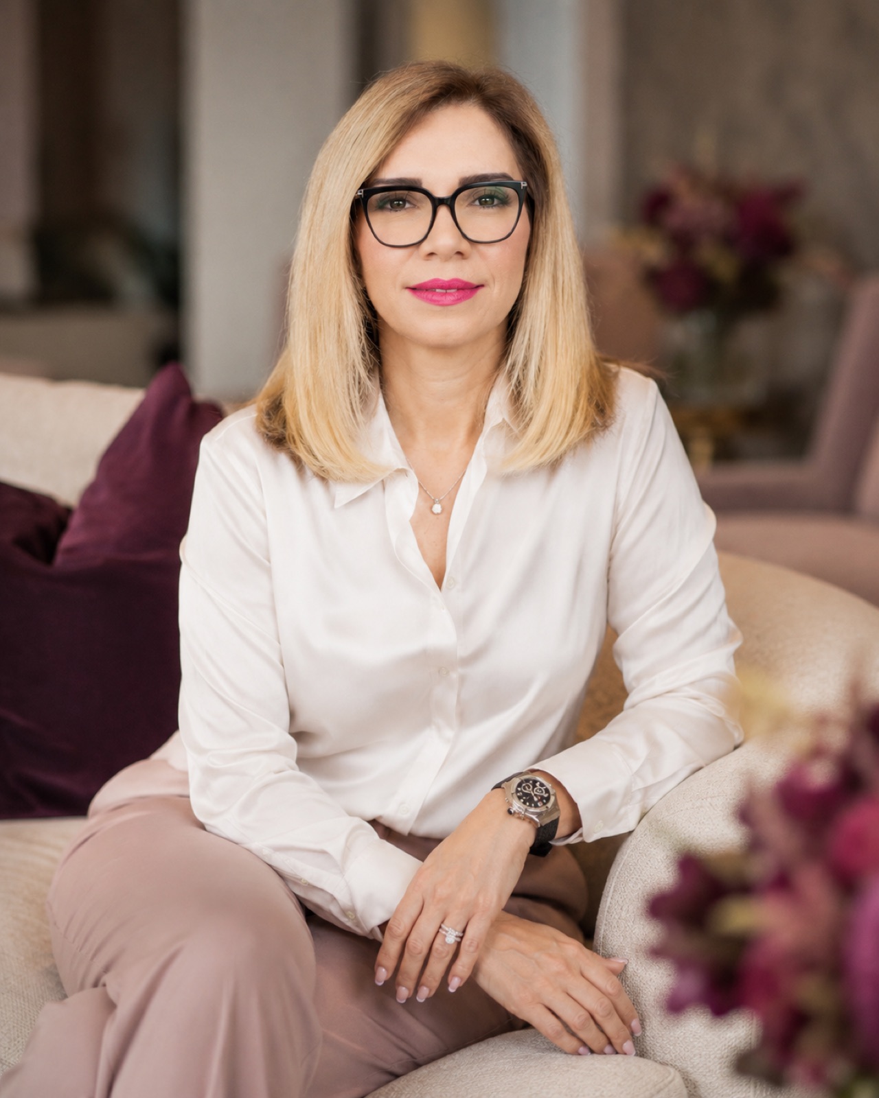 Ivory blouse, black frames. Seated in a cream sofa with plum cushions.