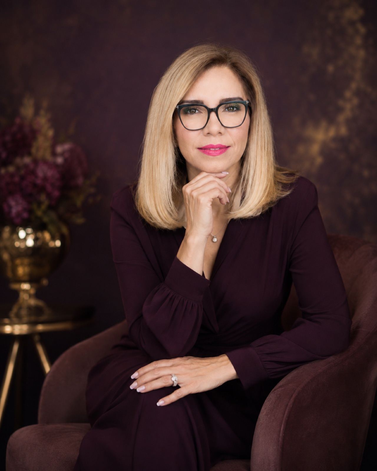 Plum dress, black frames. Hand to chin, plum ground with florals.