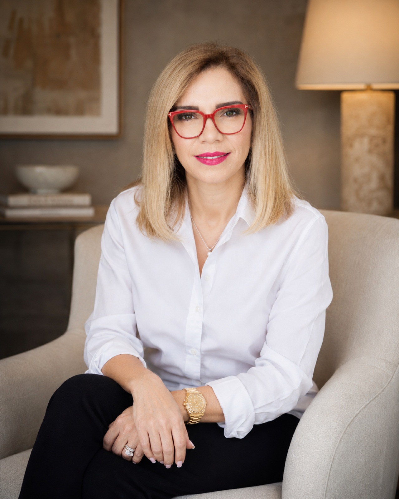 White shirt over black trousers, red frames. Seated in a cream armchair, lamplit room with artwork behind. Crops below the knee.