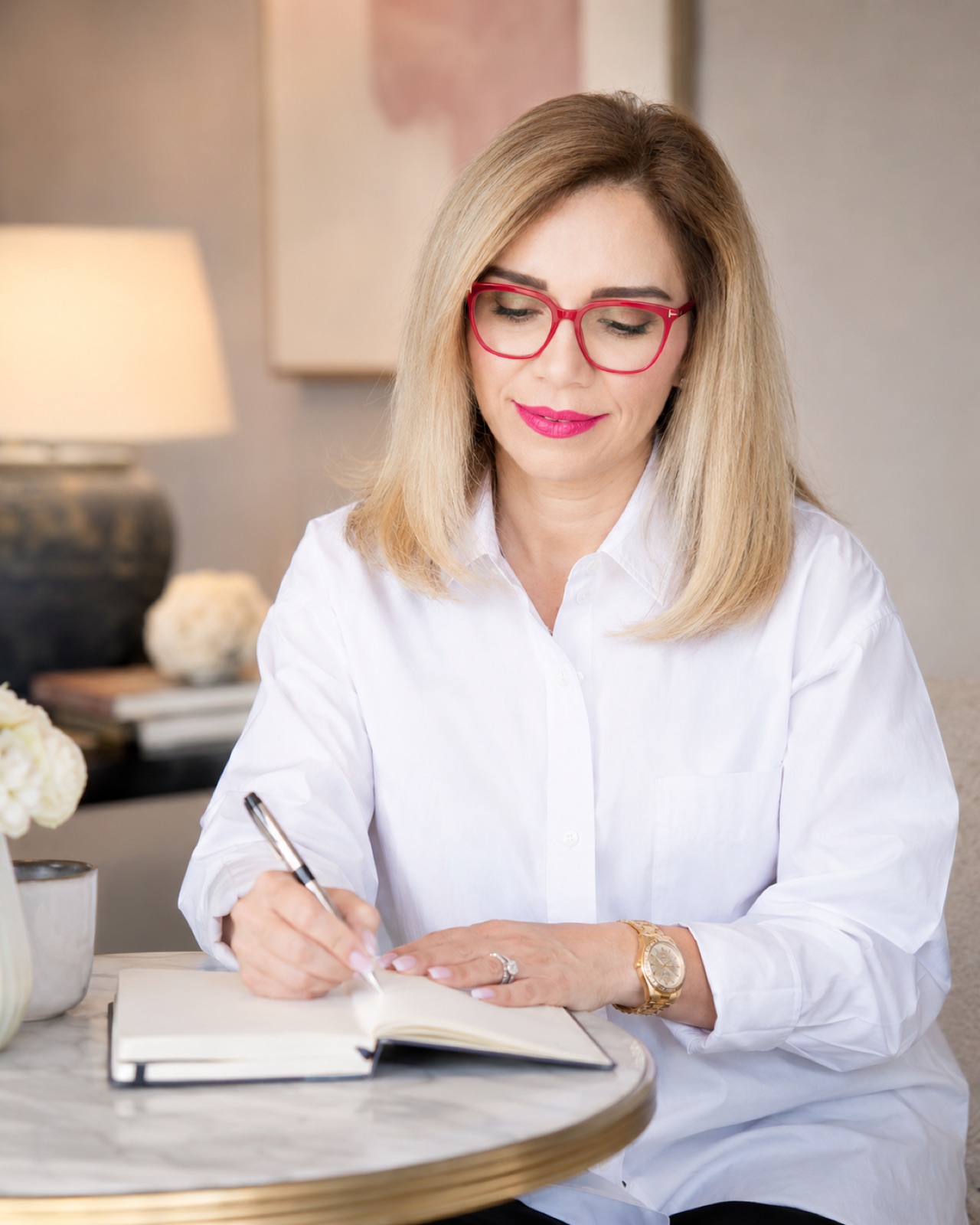 White shirt, red frames. Writing in a journal at a round marble table, roses and a lamp behind. Half body, eyes down.